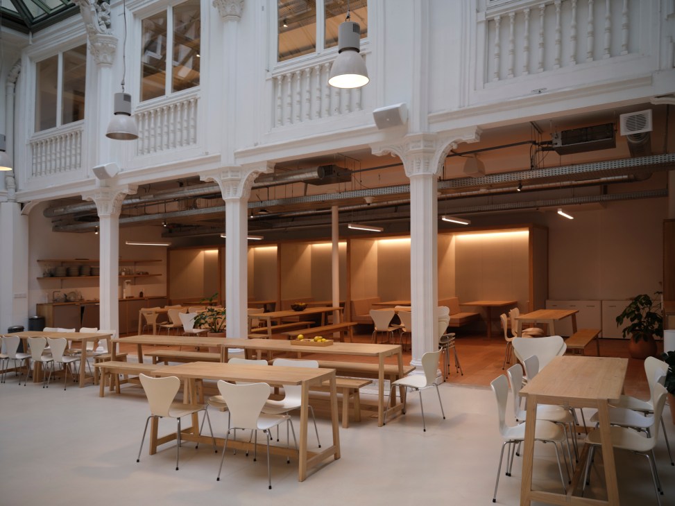Modern office dining area with wooden tables, white chairs, exposed white columns and warm lighting in historic building