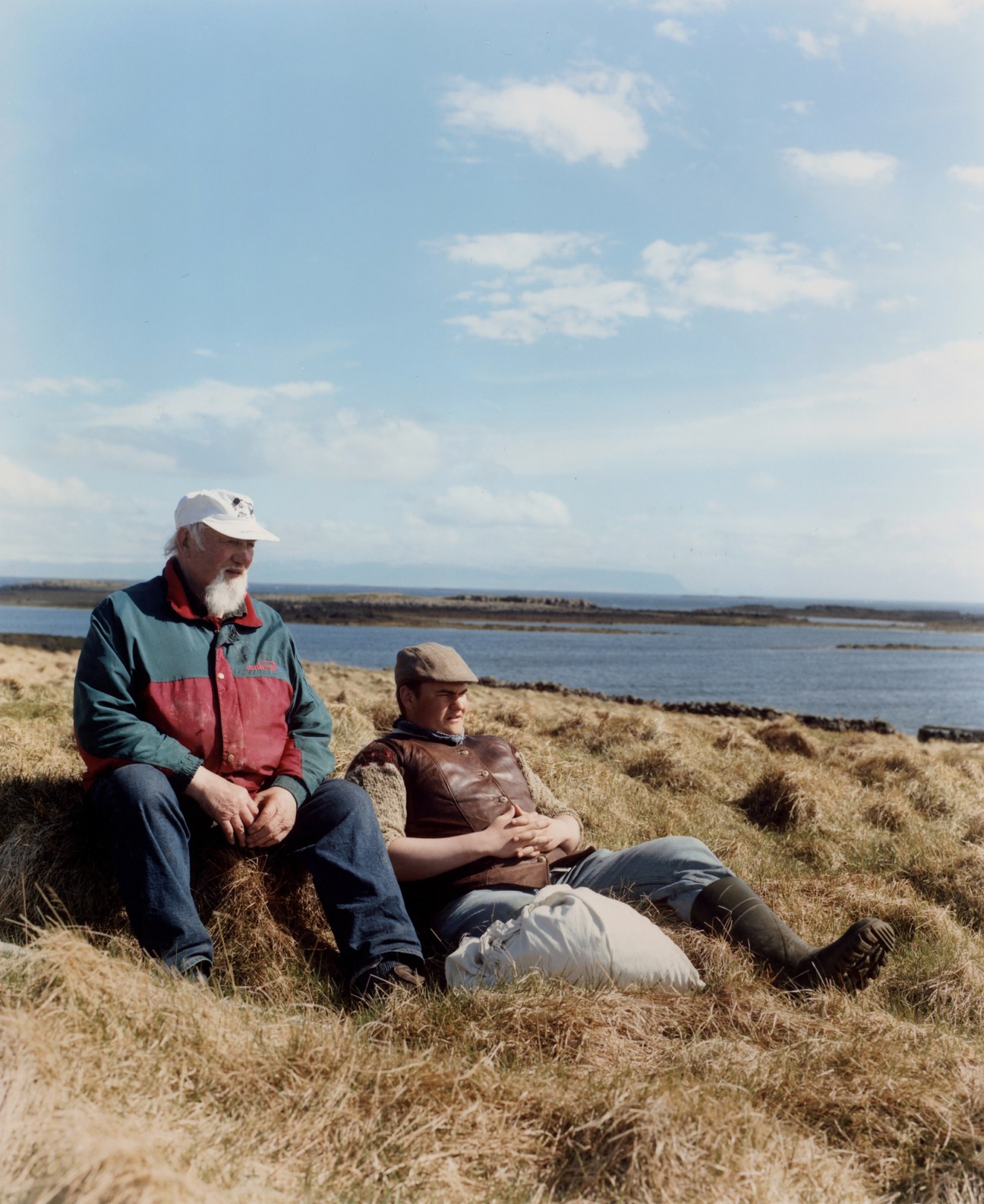 Two men in caps and jackets sitting on golden grass overlooking a coastal landscape with water and islands