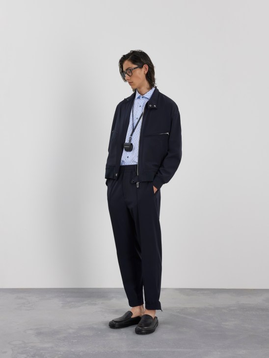 Man in navy blazer and trousers with patterned shirt, black glasses and loafers against white background