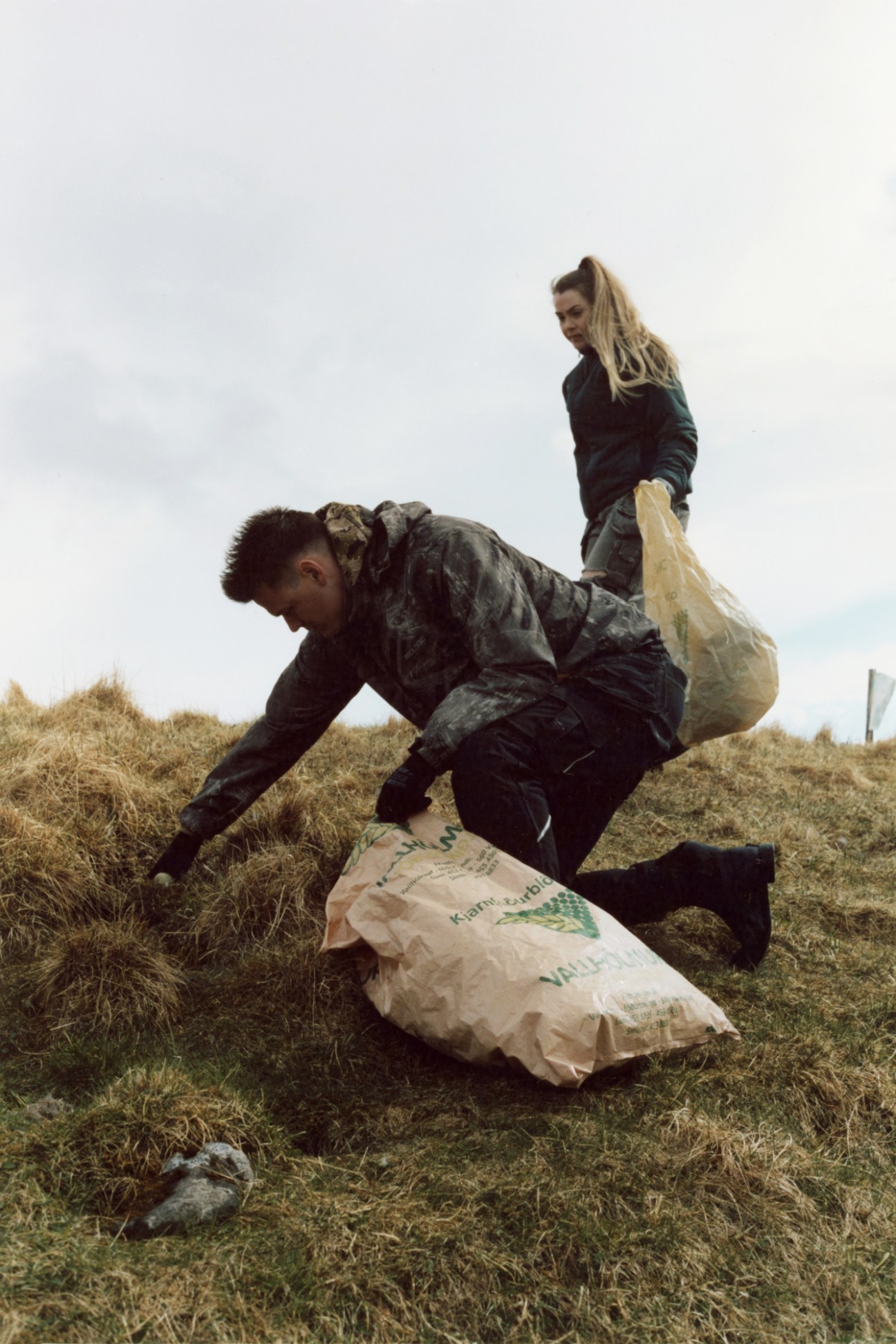 Person in camouflage clothing kneeling on grassy terrain collecting material into a large white sack