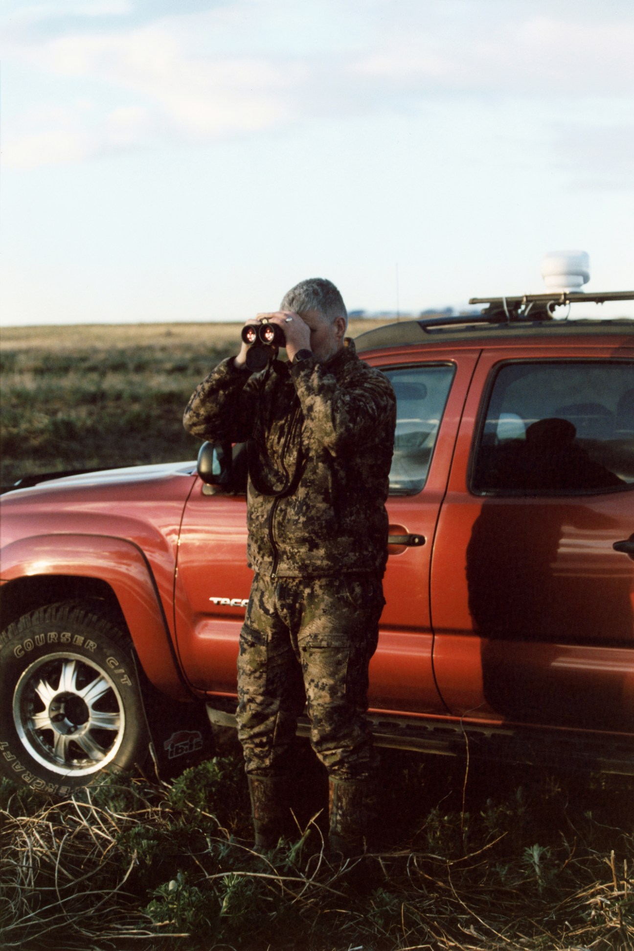 Farmer in camouflage clothing using binoculars to watch for predators near red pickup truck in Icelandic field