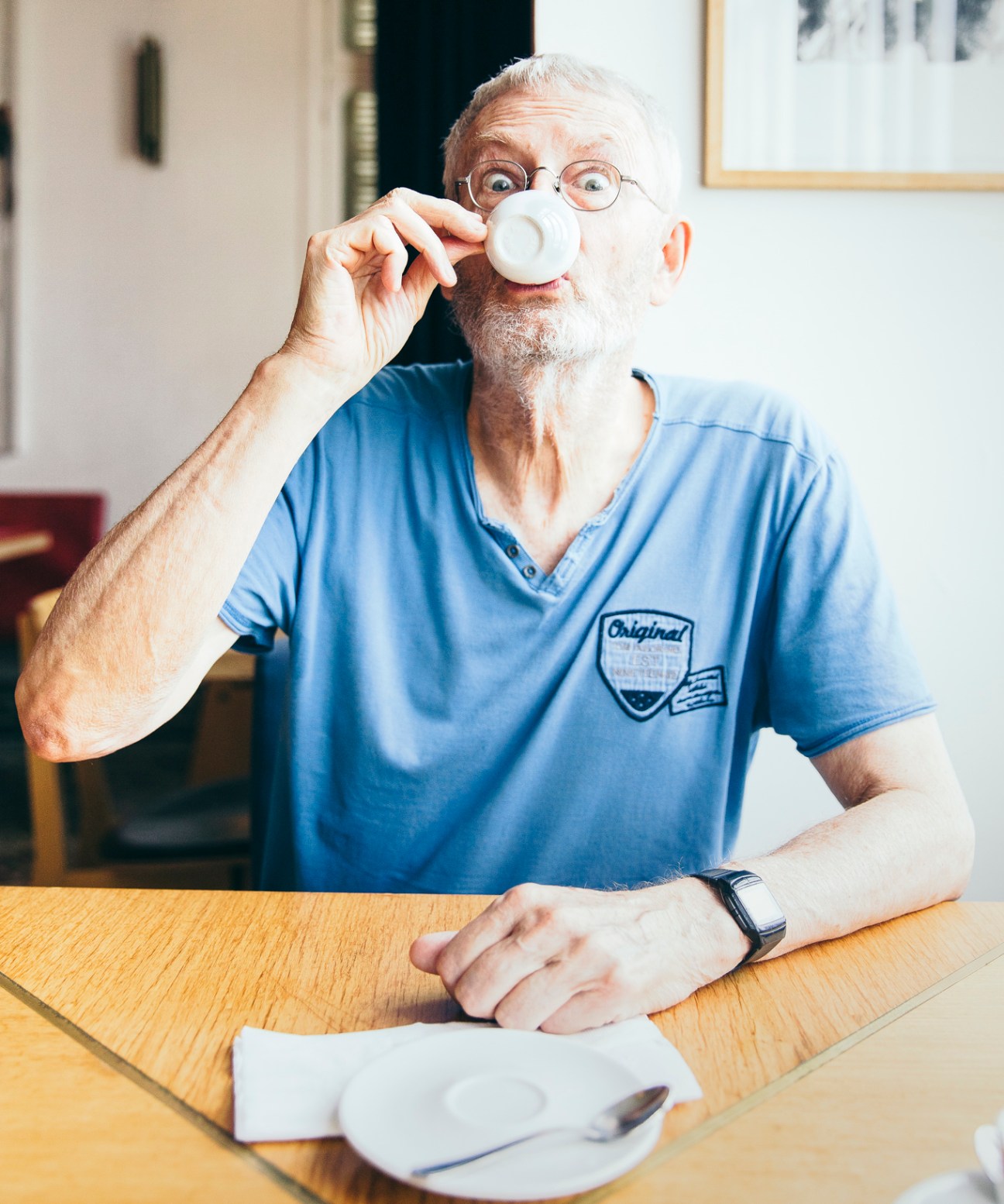 Elderly man with glasses drinking from white cup at wooden table