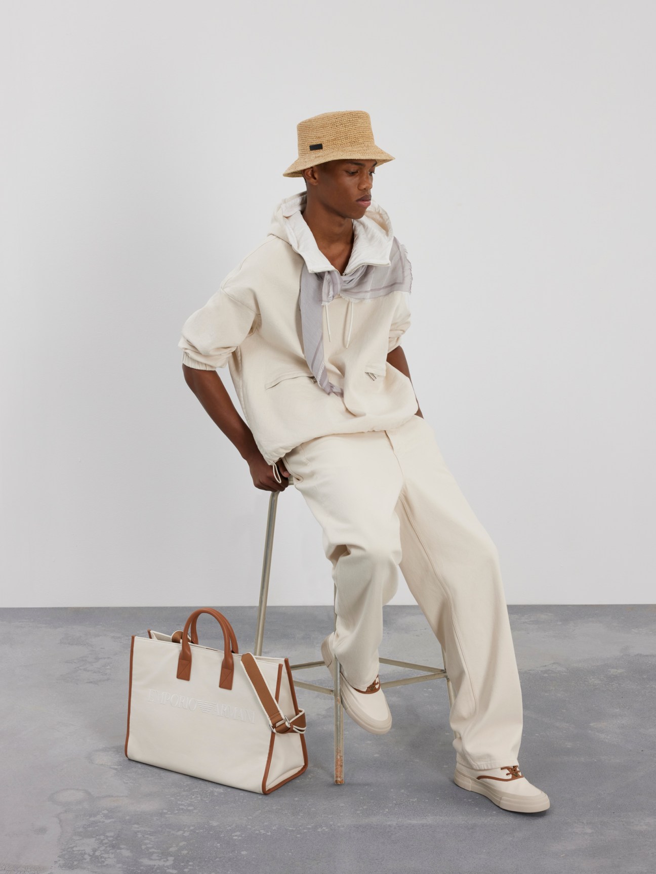 Model wearing cream-colored Emporio Armani suit with white shirt and straw bucket hat, holding a two-tone canvas and leather tote bag.