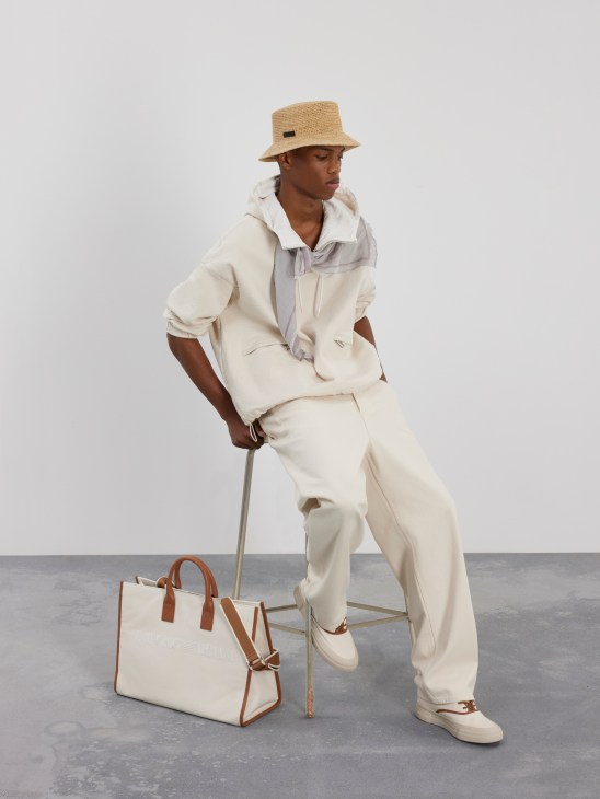 Model in cream outfit with bucket hat sitting on stool next to canvas bag in minimalist studio setting