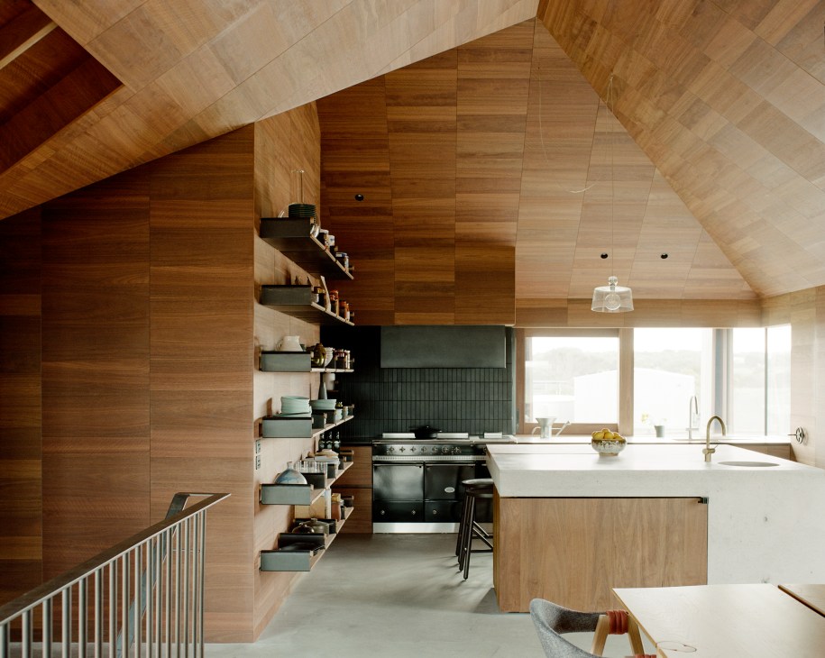 Open-plan kitchen with timber cabinetry, white island, green shelving and vaulted wood ceiling