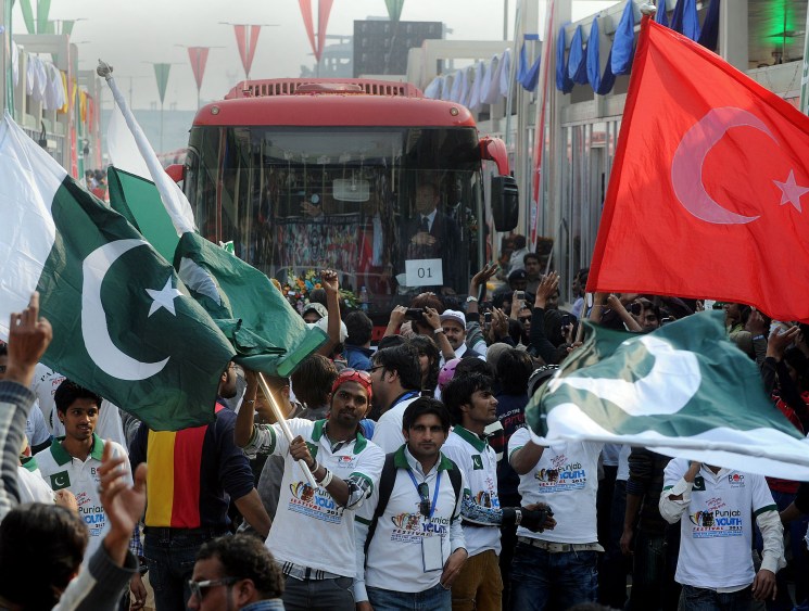 Crowd of Pakistani youth waving Pakistani and Turkish flags at a public gathering in an urban street.