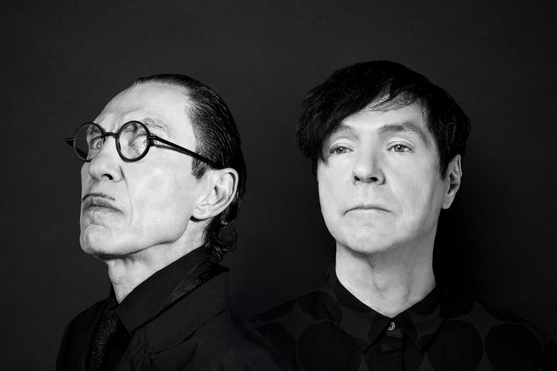 Two men in black clothing pose for a portrait against a dark background.