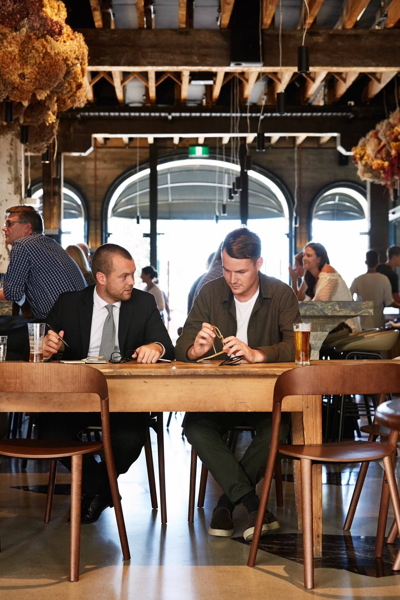 Two men sitting at a wooden bar counter, one in a checked shirt and tie, one in a casual brown jacket, discussing business over beers in a bright industrial venue.