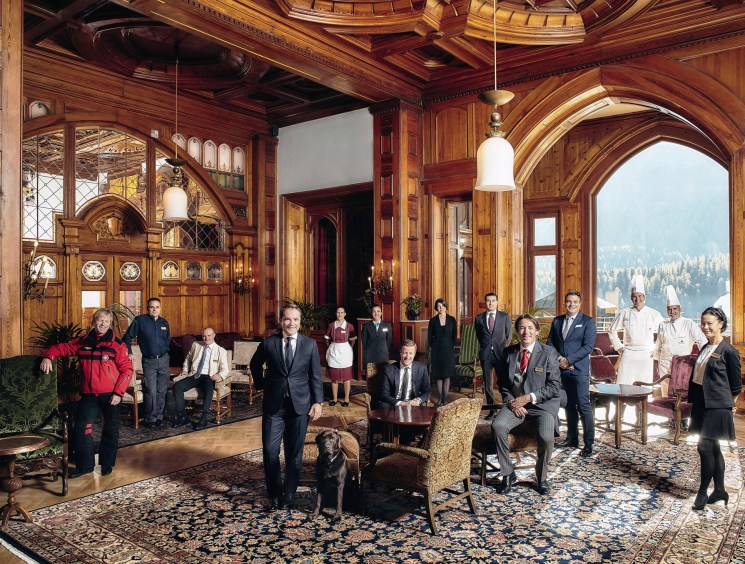 A large hotel staff group posed in an ornate wood-panelled grand hall with arched windows overlooking a lake or mountain view.