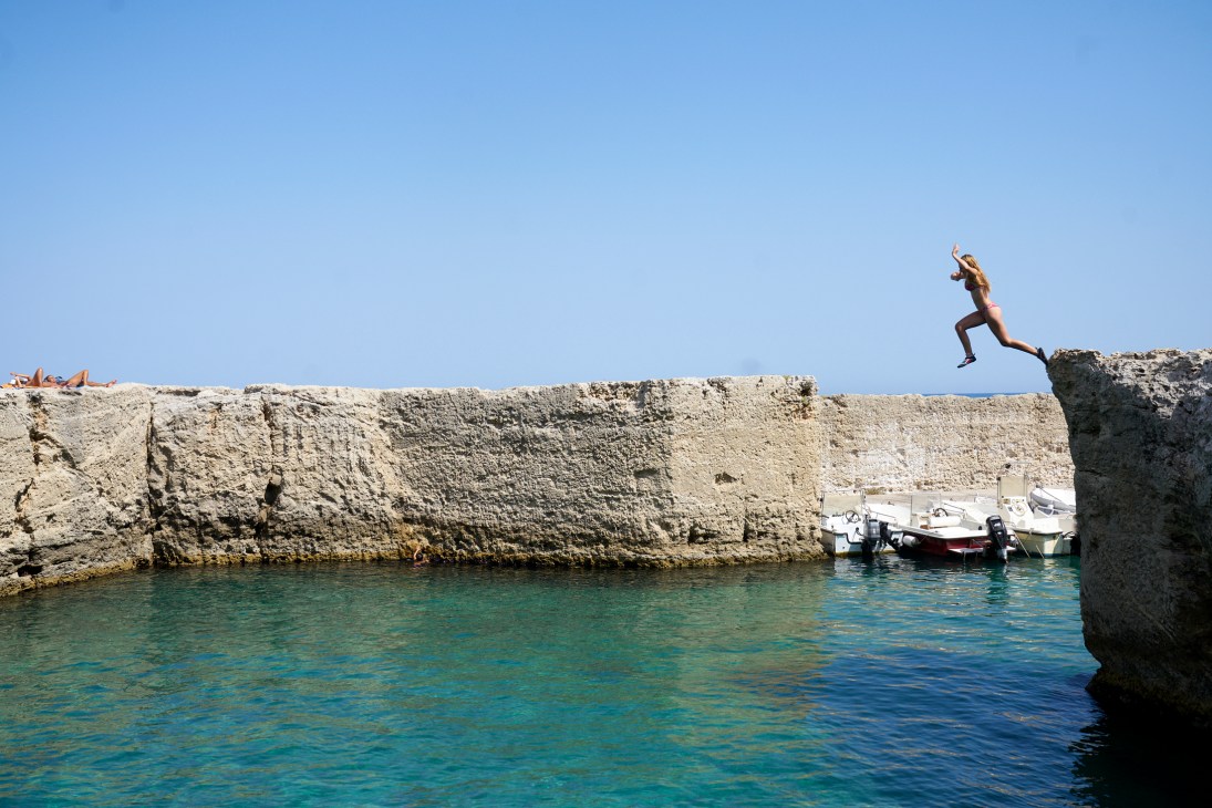 A person mid-jump from dramatic limestone cliffs into bright blue water below