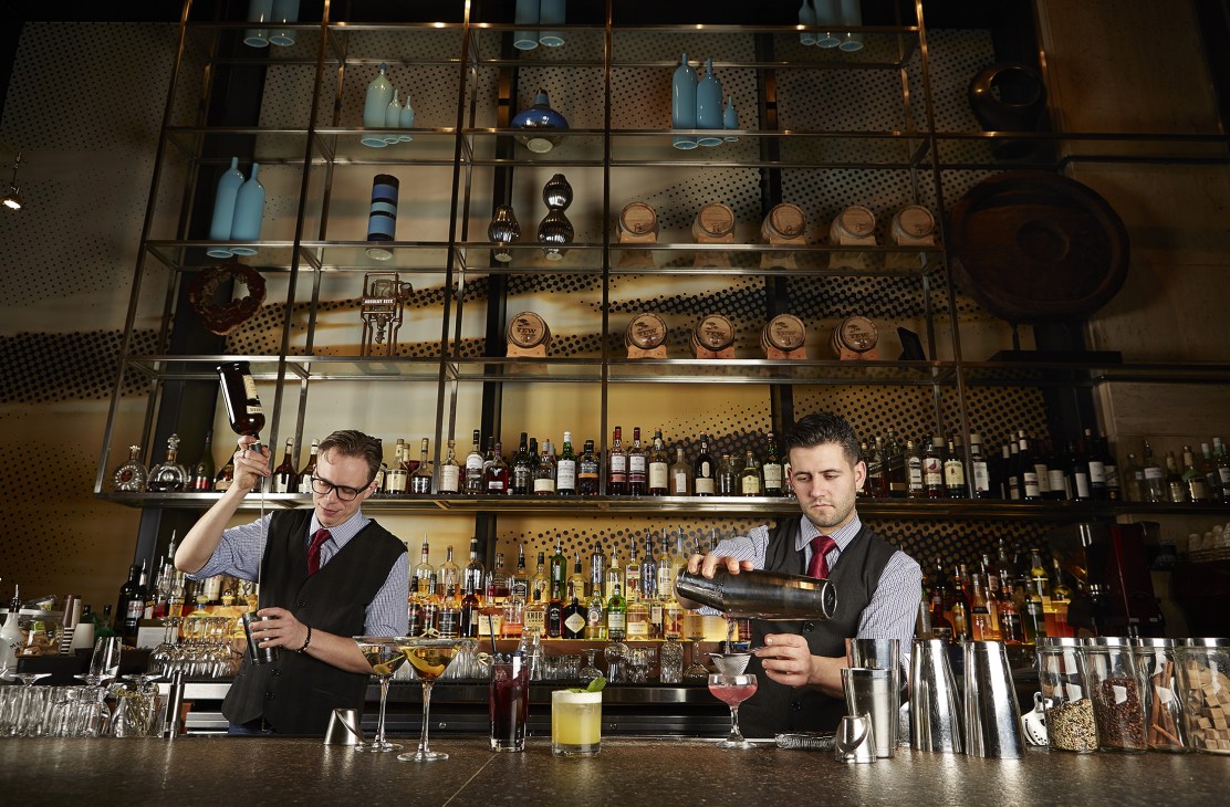 Two bartenders prepare cocktails behind a counter at an upscale bar with shelves of spirits and decorative bottles.