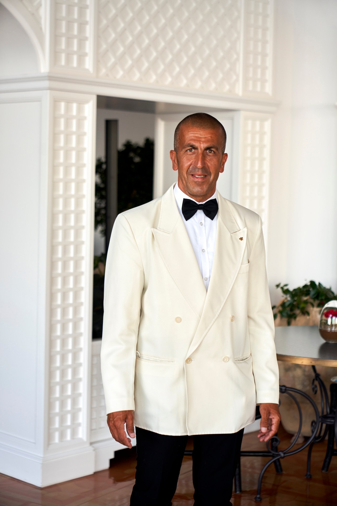 Man in cream double-breasted jacket and black bow tie standing in white Mediterranean-style interior