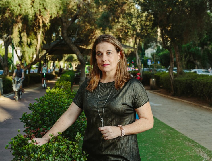 A woman in a dark dress stands on a tree-lined street in Tel Aviv, with one hand resting on green shrubs