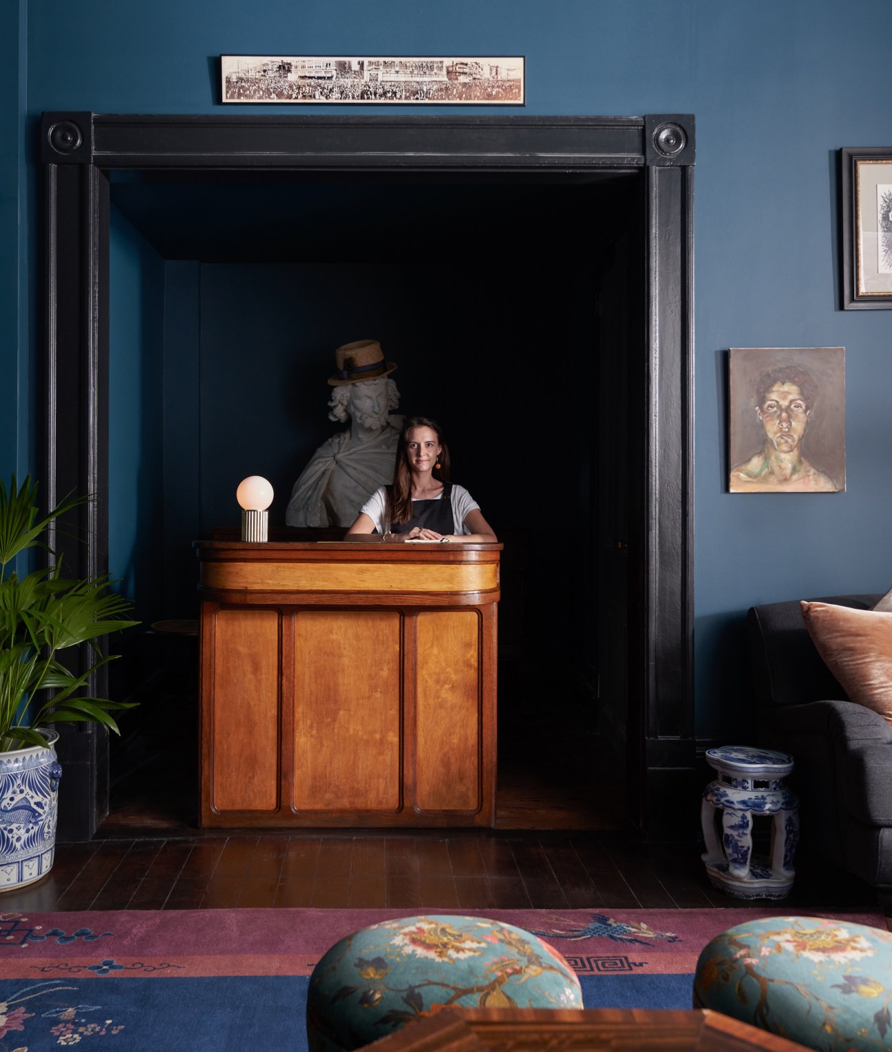 Hotel reception desk with female staff member behind wooden counter in blue-walled interior with artwork