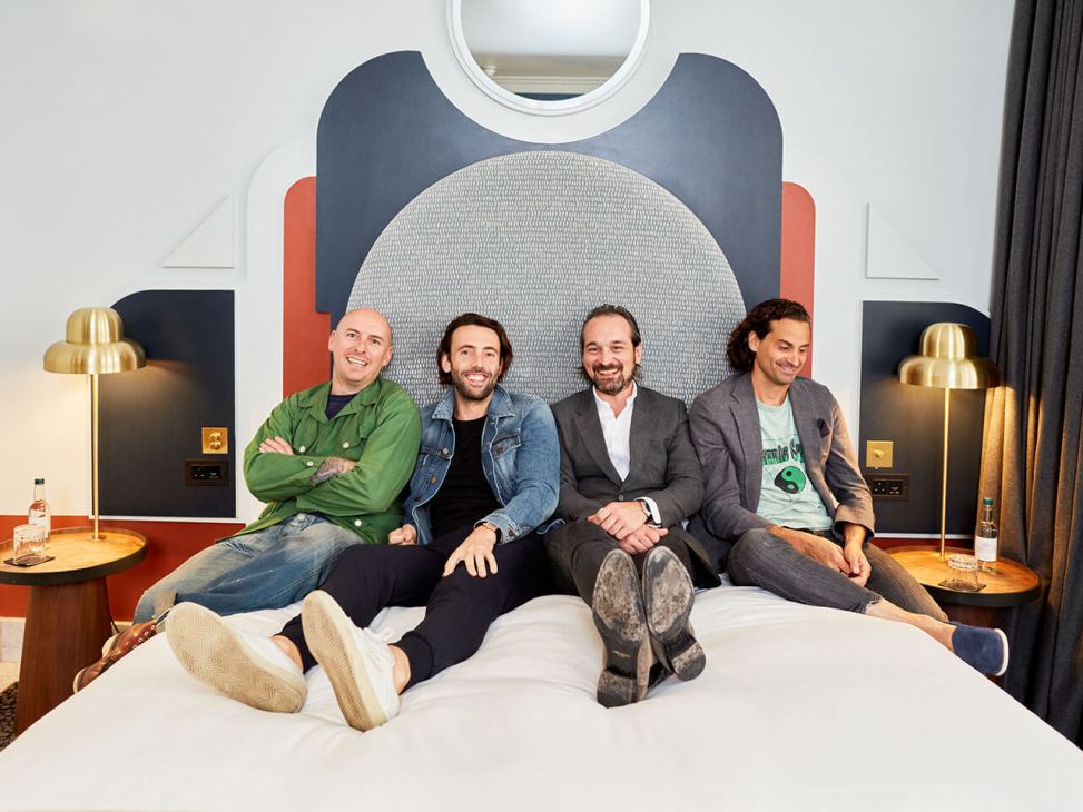 Four men sitting on a hotel bed smiling at camera in modern room with colorful headboard