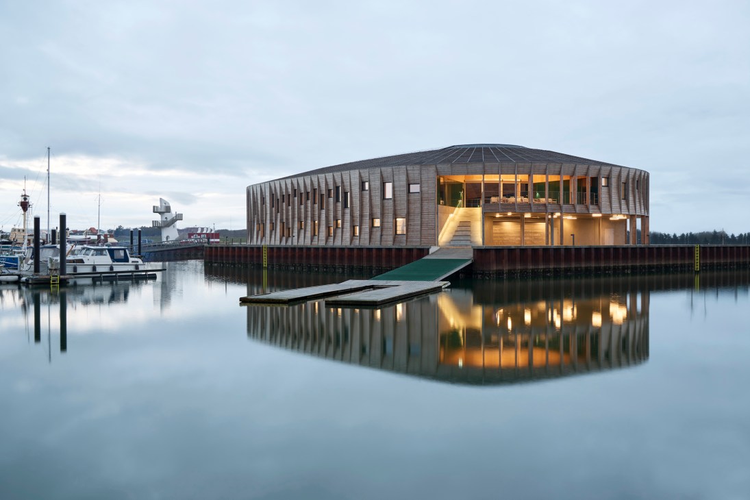Circular modern building on water with wooden facade and glass windows reflecting in calm harbor