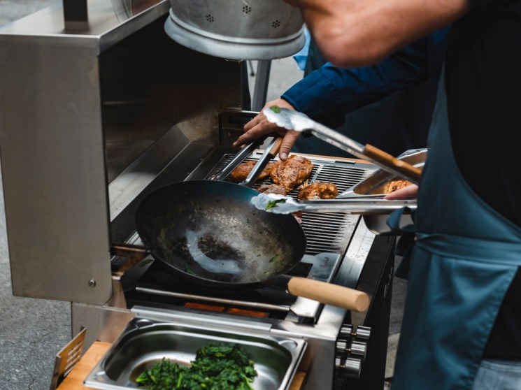 Person operating a Flammkraft grill, handling leafy greens in a colander over the cooking surface