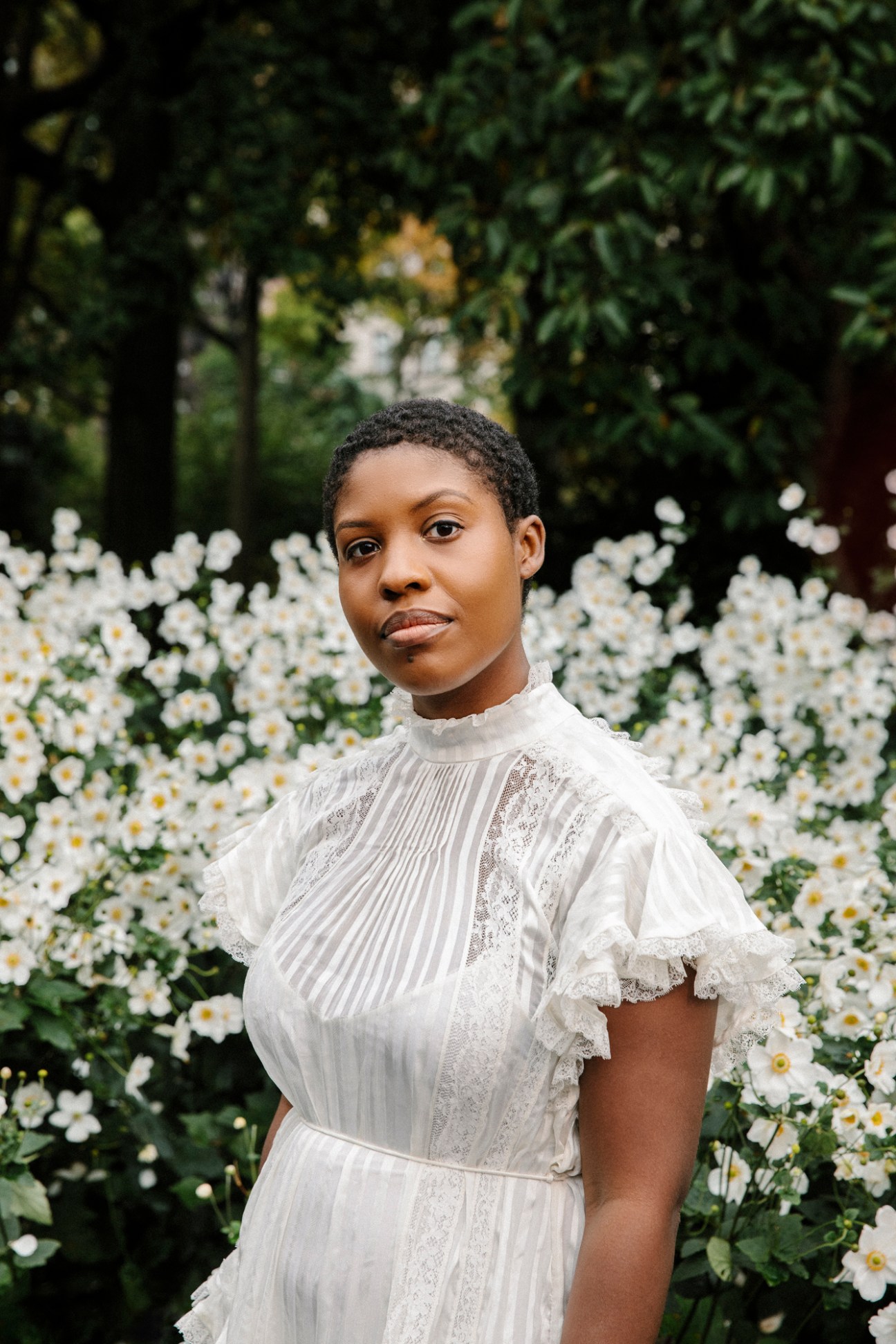Portrait of author Raven Leilani wearing a white lace dress against a floral background