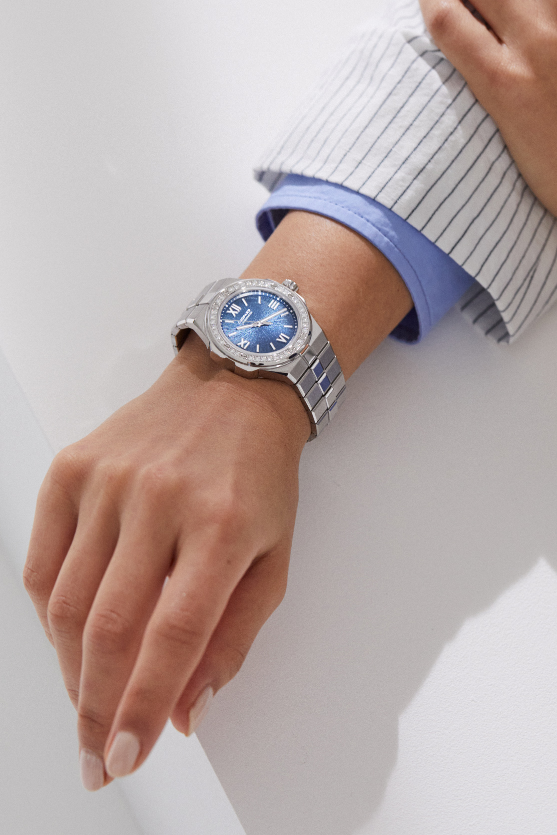 A hand wearing a Chopard Alpine Eagle watch with blue dial and diamond bezel, dressed in a striped shirt and blue jacket