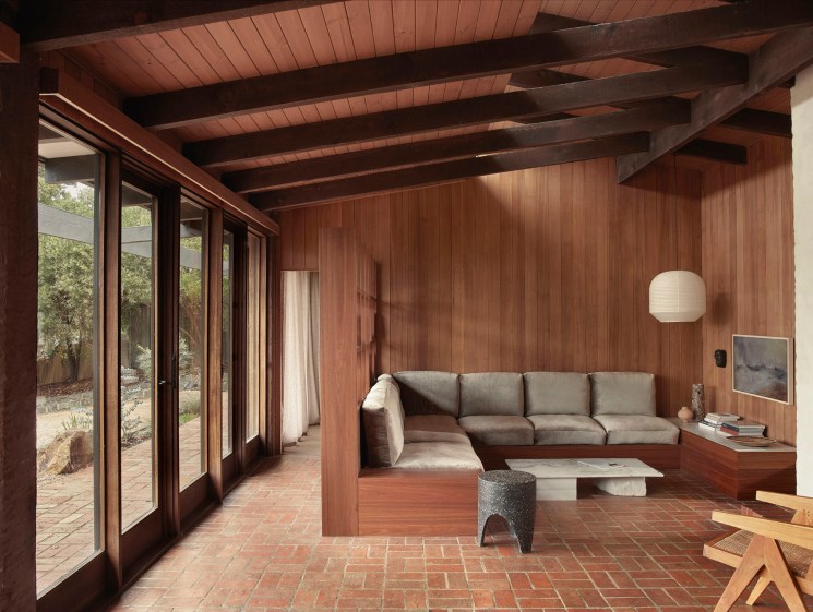Modernist living room with timber-lined built-in sofa, brick floors, and large glass doors opening to garden.