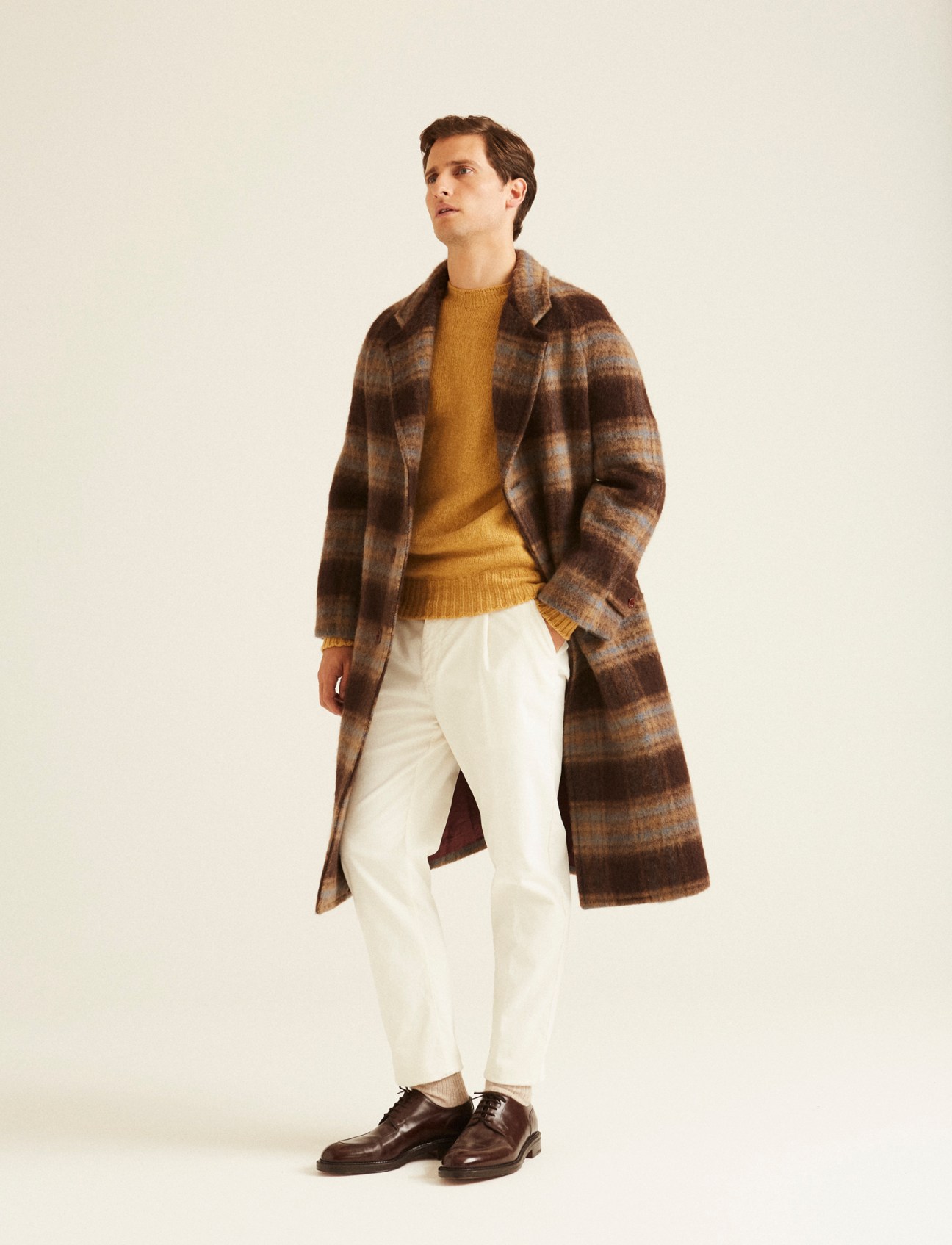 Man in brown striped wool coat over mustard sweater and white trousers, looking away from camera