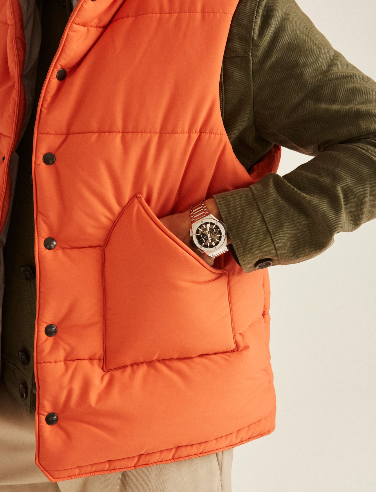 Person wearing olive green jacket with orange quilted vest lining and luxury watch, hand in pocket