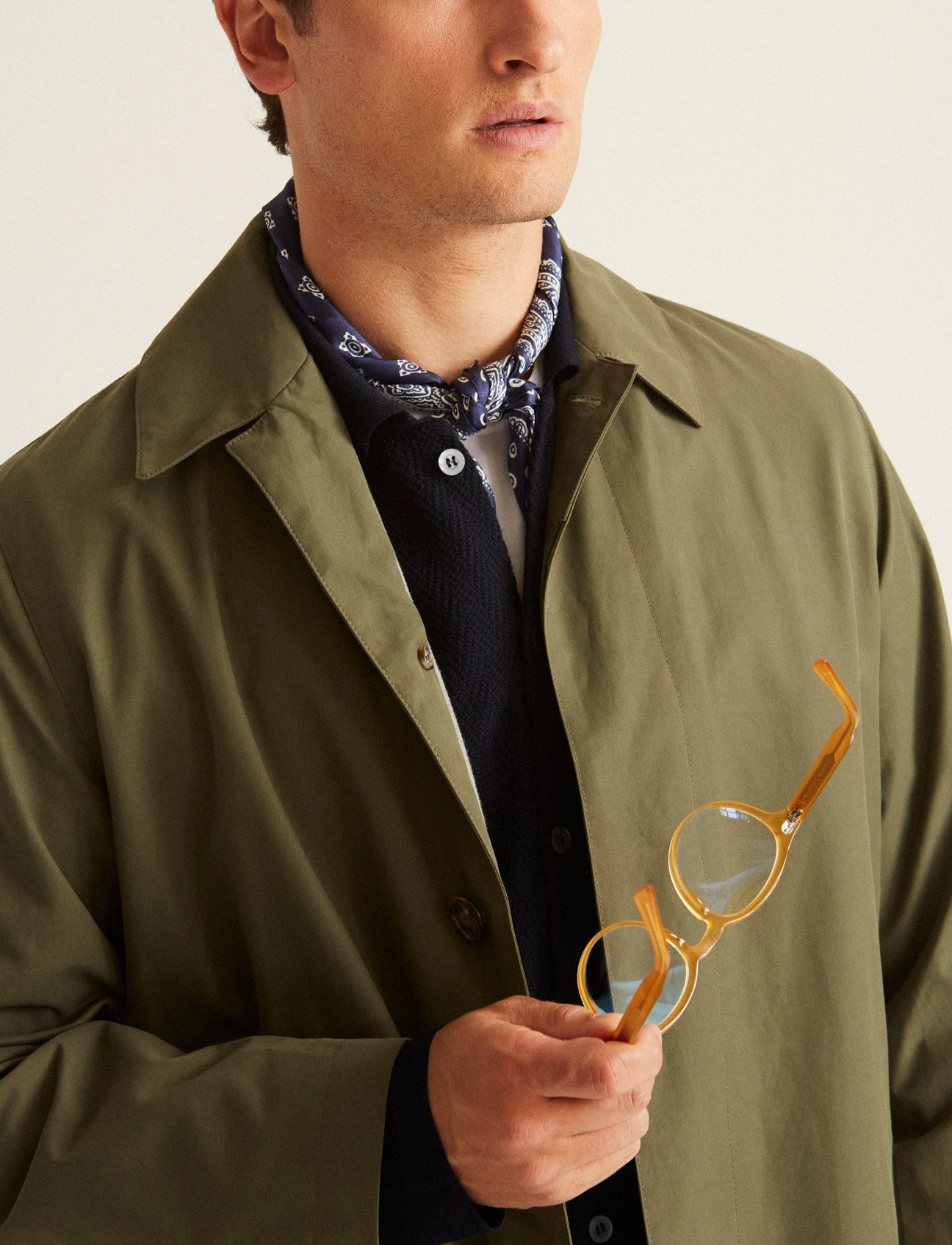 Man in olive green trench coat over dark shirt with bandana, holding round sunglasses with wooden arms