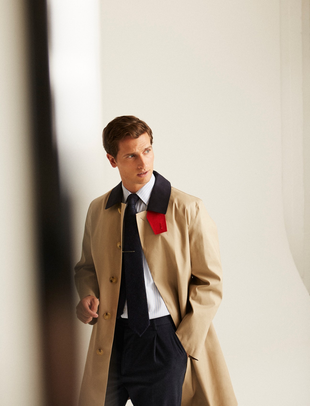 Man in beige trench coat with dark collar over navy sweater and tie against neutral background