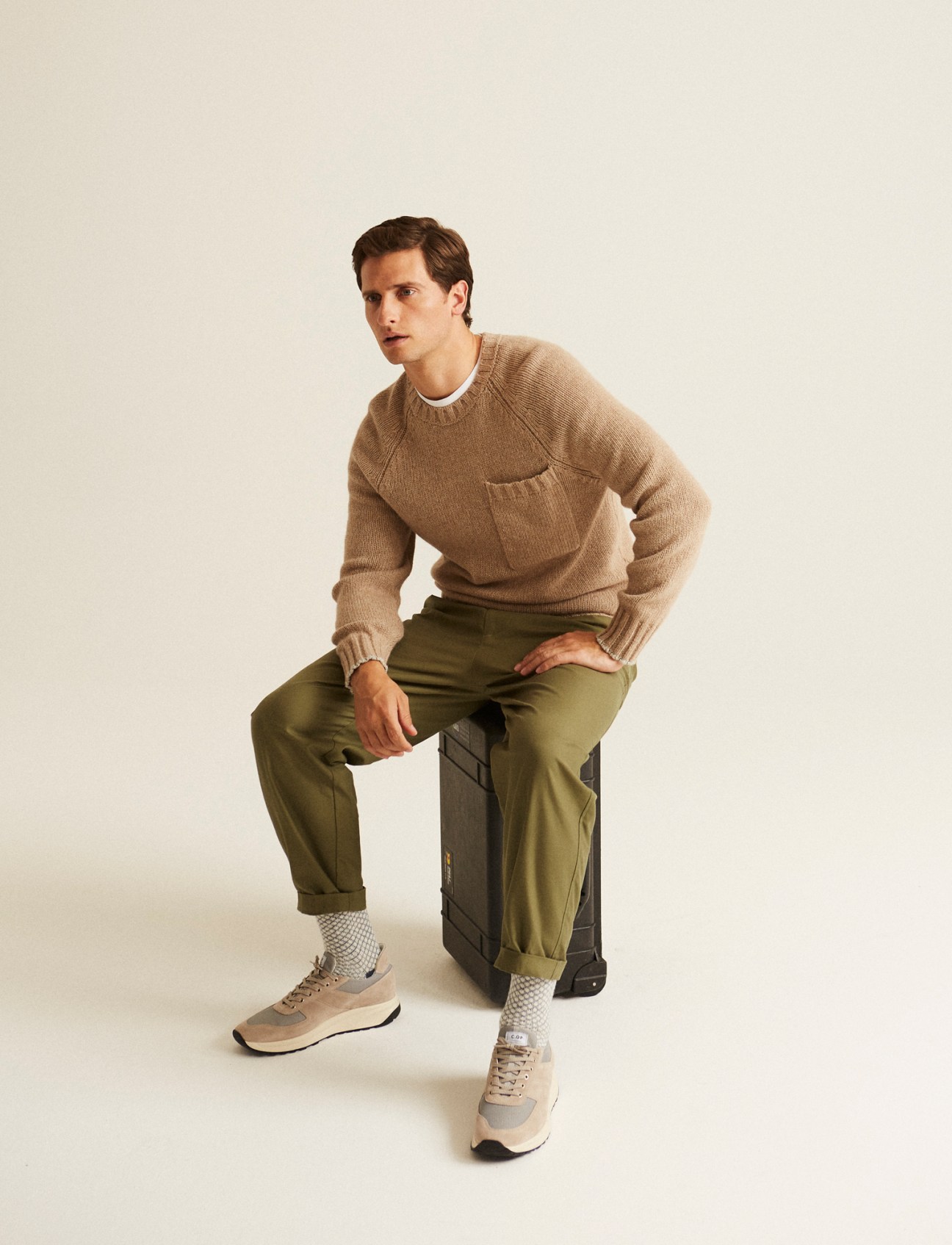 Man in beige knit sweater and olive green trousers sitting on dark suitcase wearing neutral sneakers