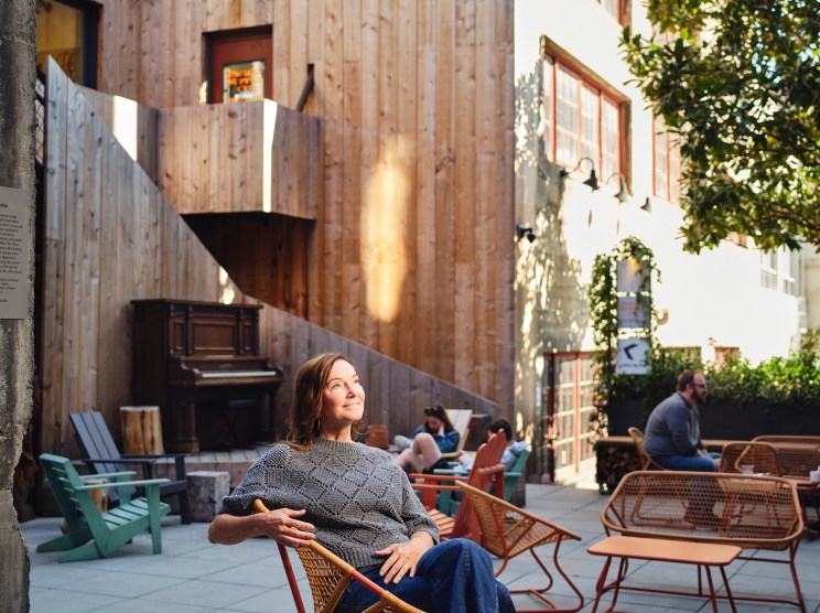Woman sitting in a modern chair in a sunlit courtyard between renovated buildings with wooden facade.