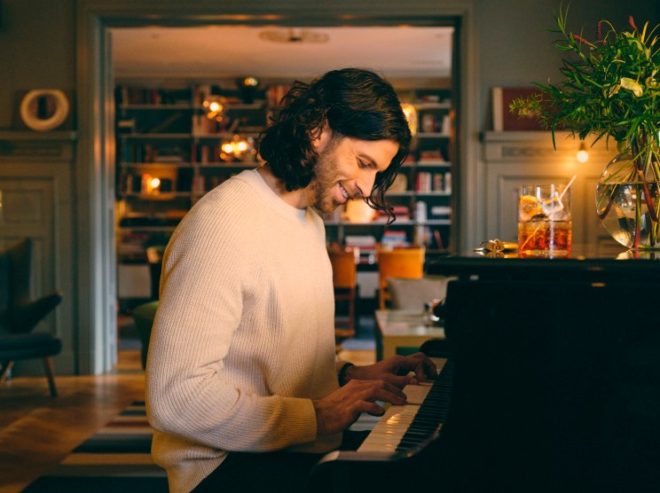 Man in cream sweater playing piano in cozy living room with bookshelves and warm lighting