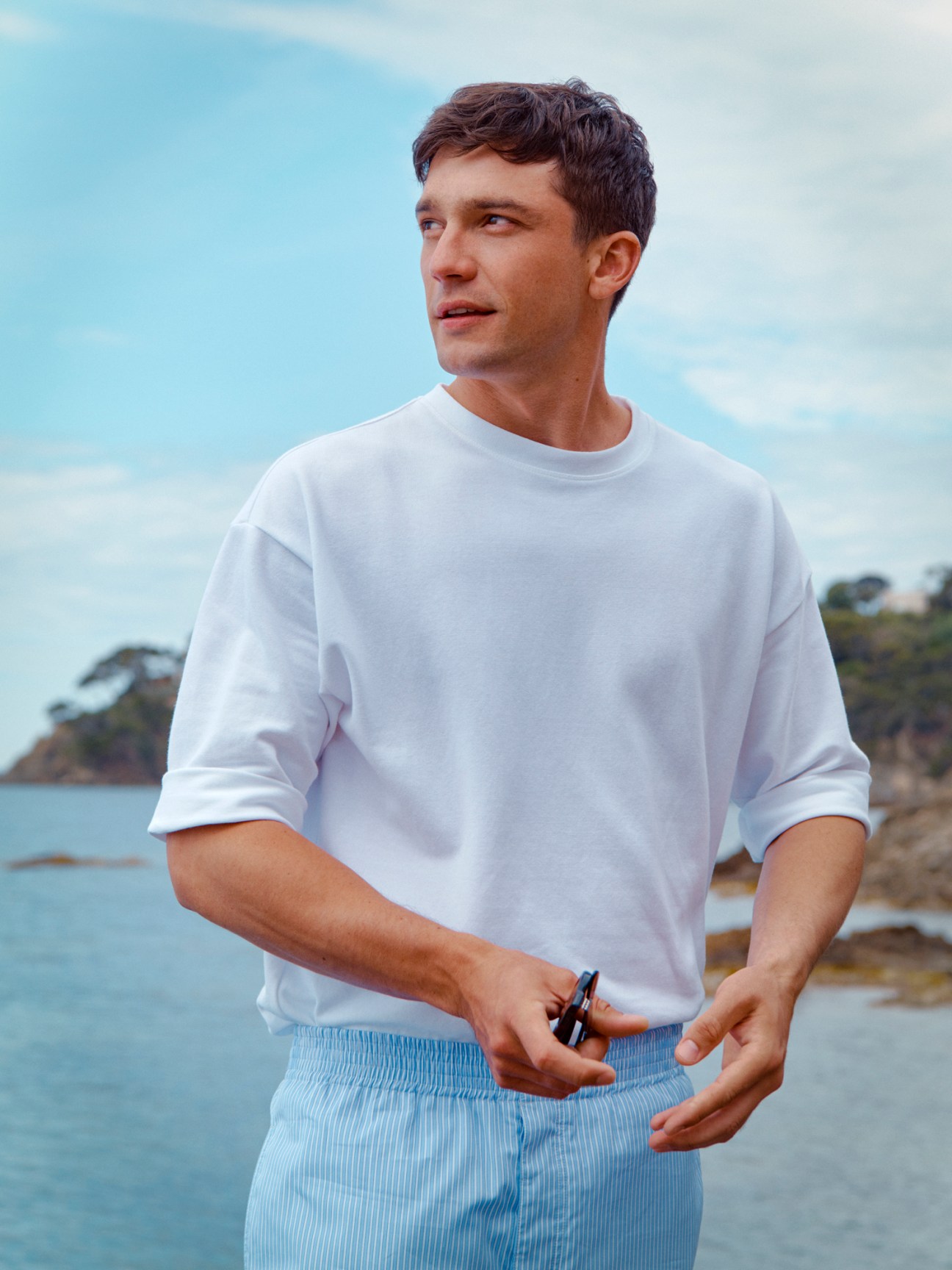 Man wearing white t-shirt and light blue striped trousers by Hermès standing by the sea.
