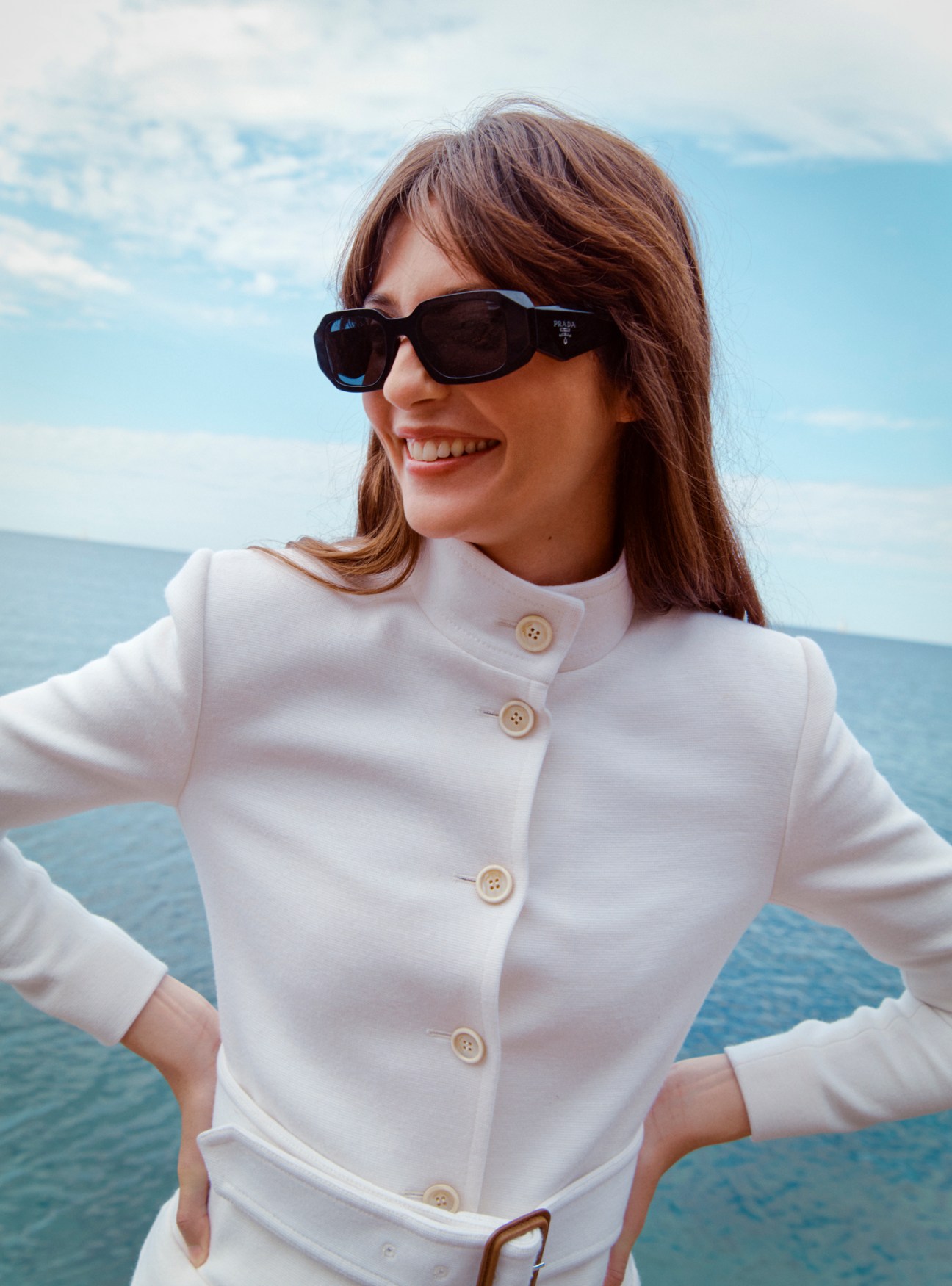 Model wearing white Saint Laurent dress and Prada sunglasses by the sea.