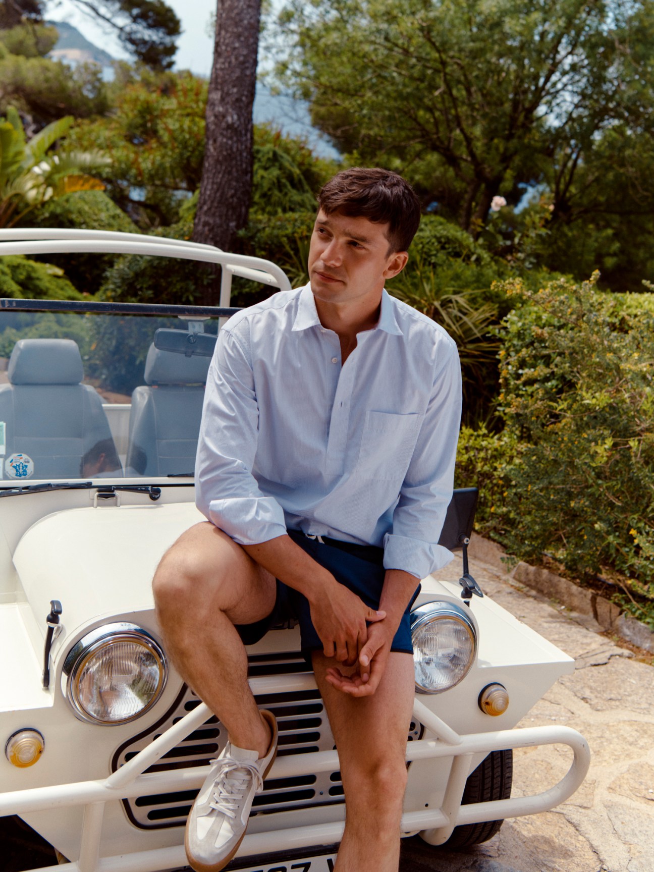 Man in light blue shirt and navy shorts sitting on white vintage jeep in Mediterranean garden setting.