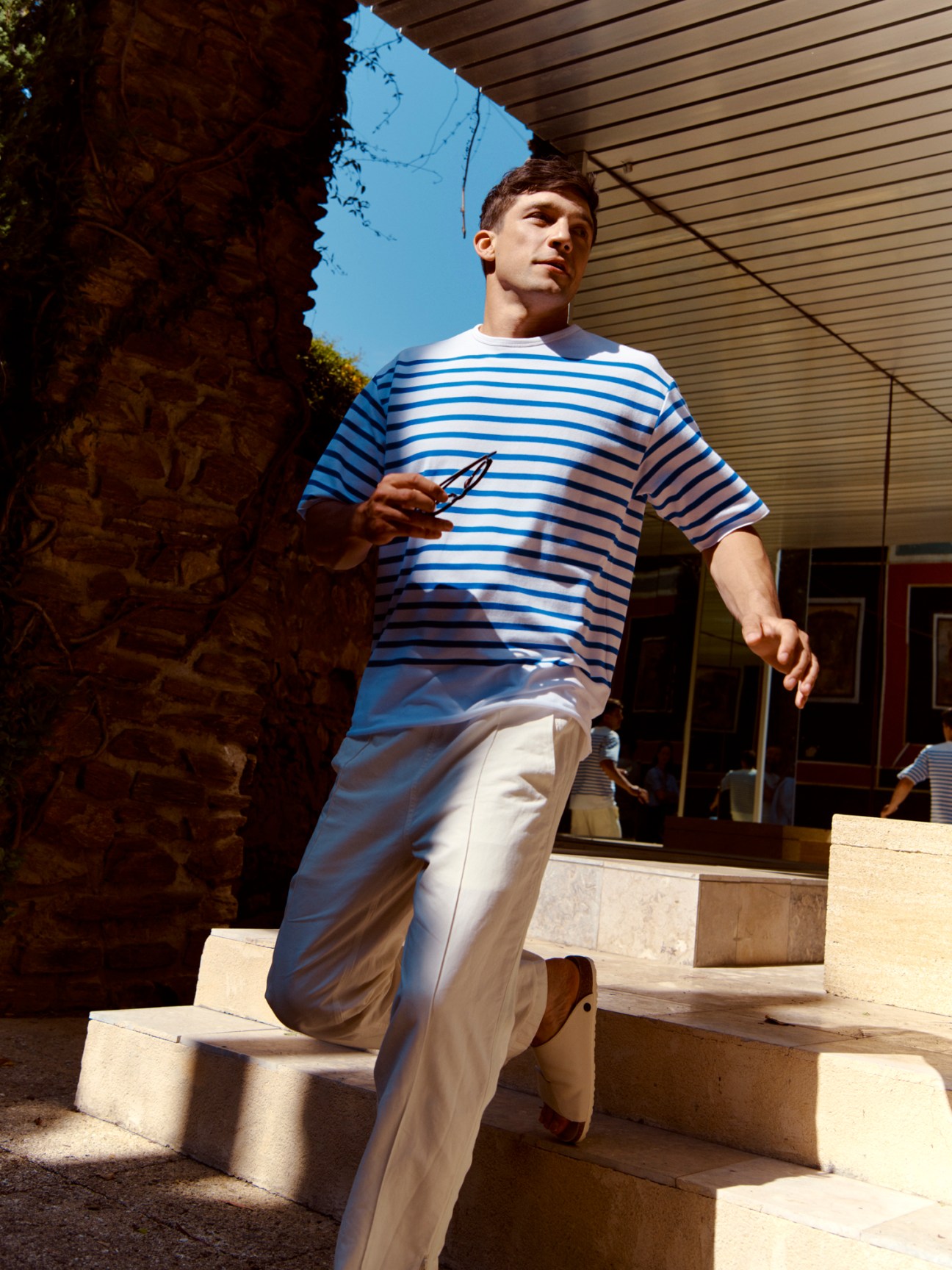 Man wearing blue and white striped t-shirt by Arpenteur and cream trousers in sunlit courtyard