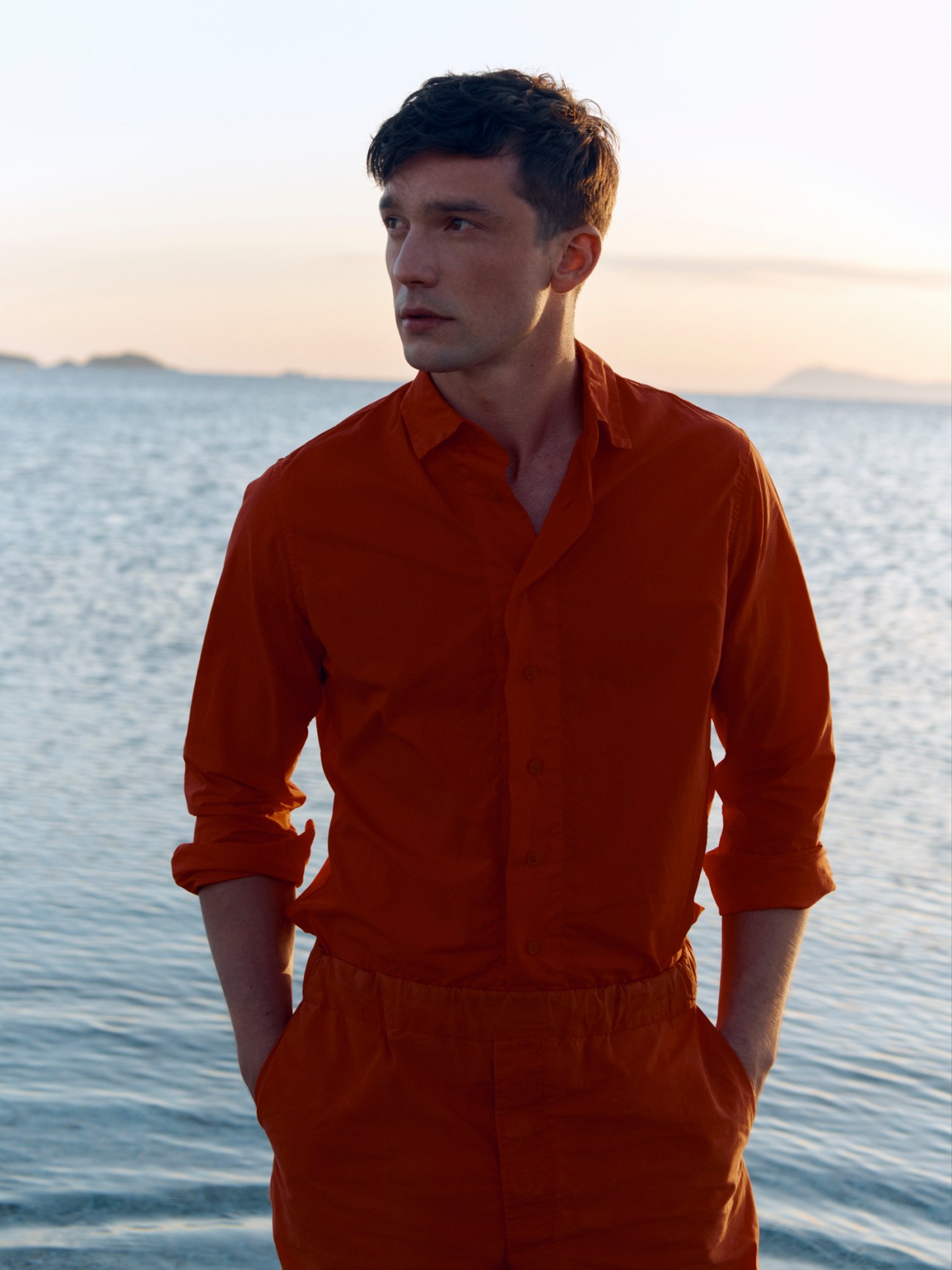 Man wearing rust-orange shirt and trousers by Casey Casey standing by the sea at sunset.