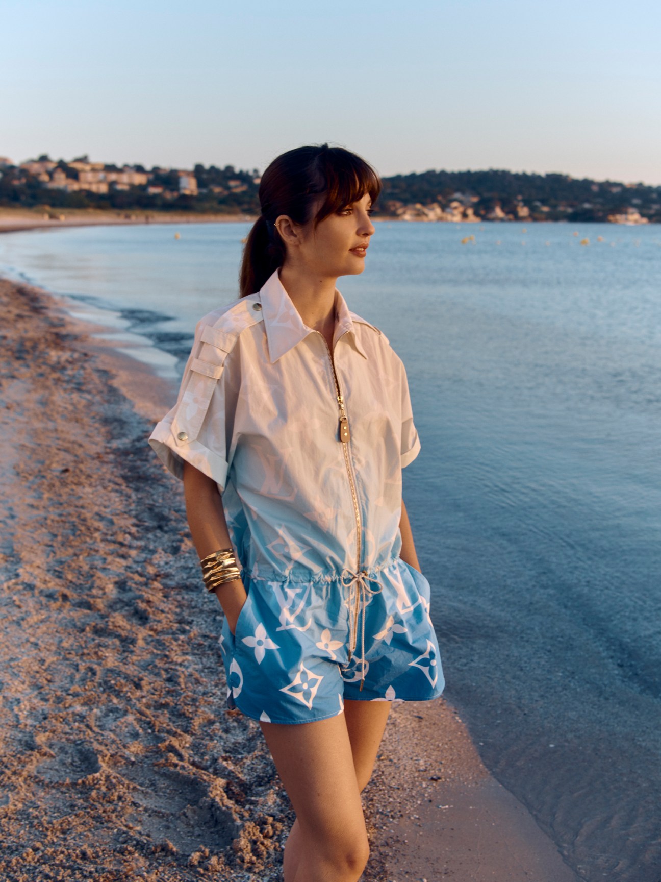 Woman in Louis Vuitton playsuit and Annelise Michelson bracelet standing on beach at sunset.