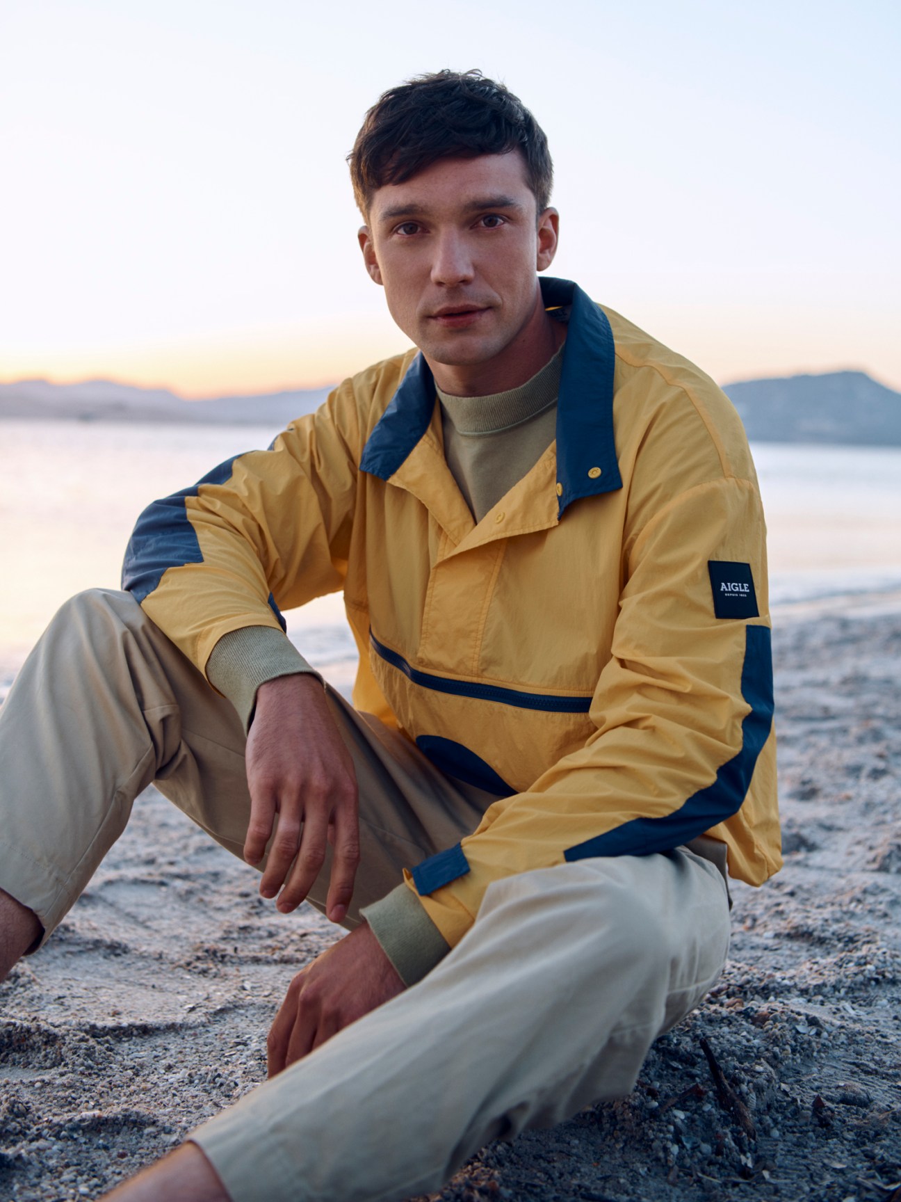 Man wearing yellow Aigle windbreaker, Margaret Howell t-shirt and Casey Casey trousers sitting on beach at sunset.