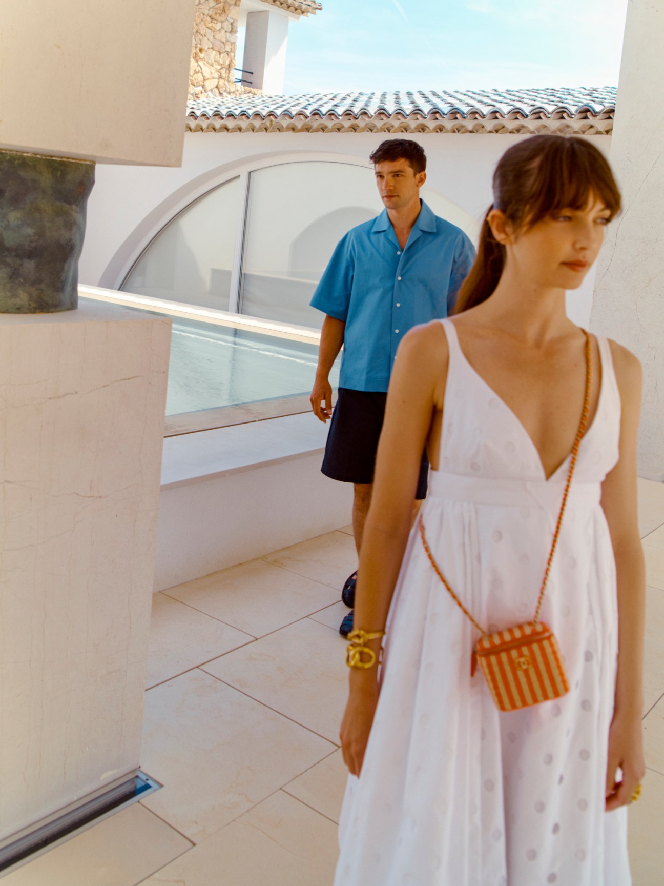 Woman in white Prada dress with orange Chanel bag and man in blue Uniforme shirt at modern Mediterranean villa.