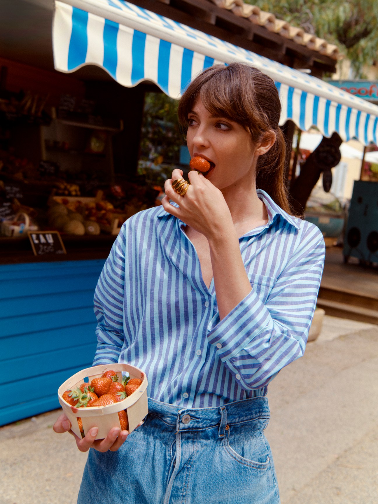 Model wearing blue striped blouse and jeans eating strawberries at a Riviera market stall.