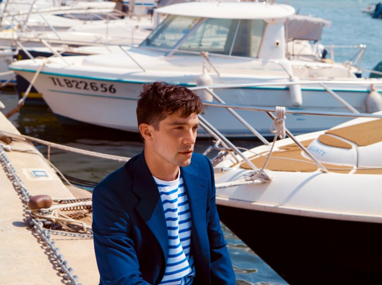 Man in blue Louis Vuitton jacket and trousers with Arpenteur striped t-shirt sitting on a dock surrounded by moored boats.