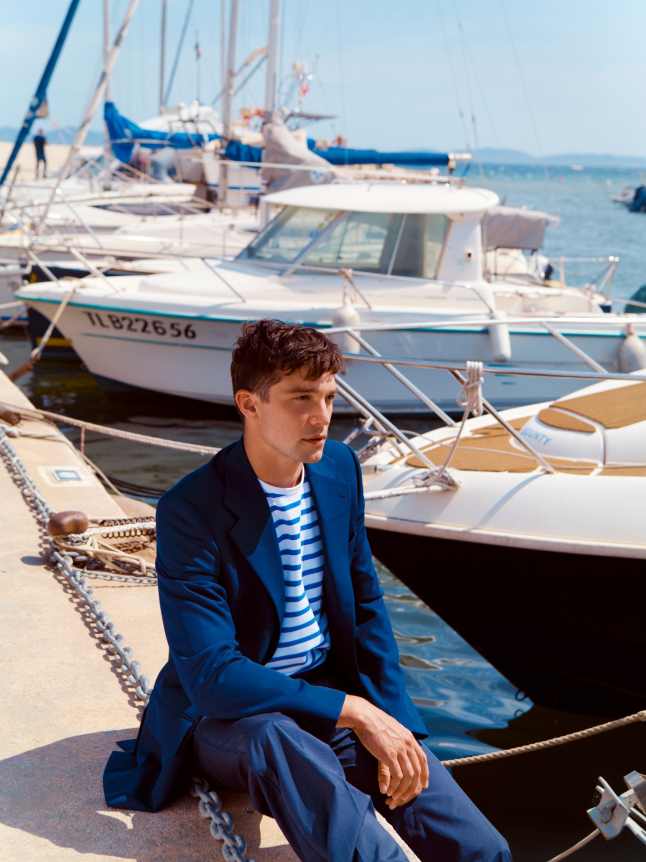 Man in blue Louis Vuitton jacket and trousers with Arpenteur striped t-shirt sitting on a dock surrounded by moored boats.