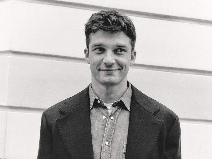 Model wearing dark blazer over light button-up shirt with hands in pockets against white paneled wall