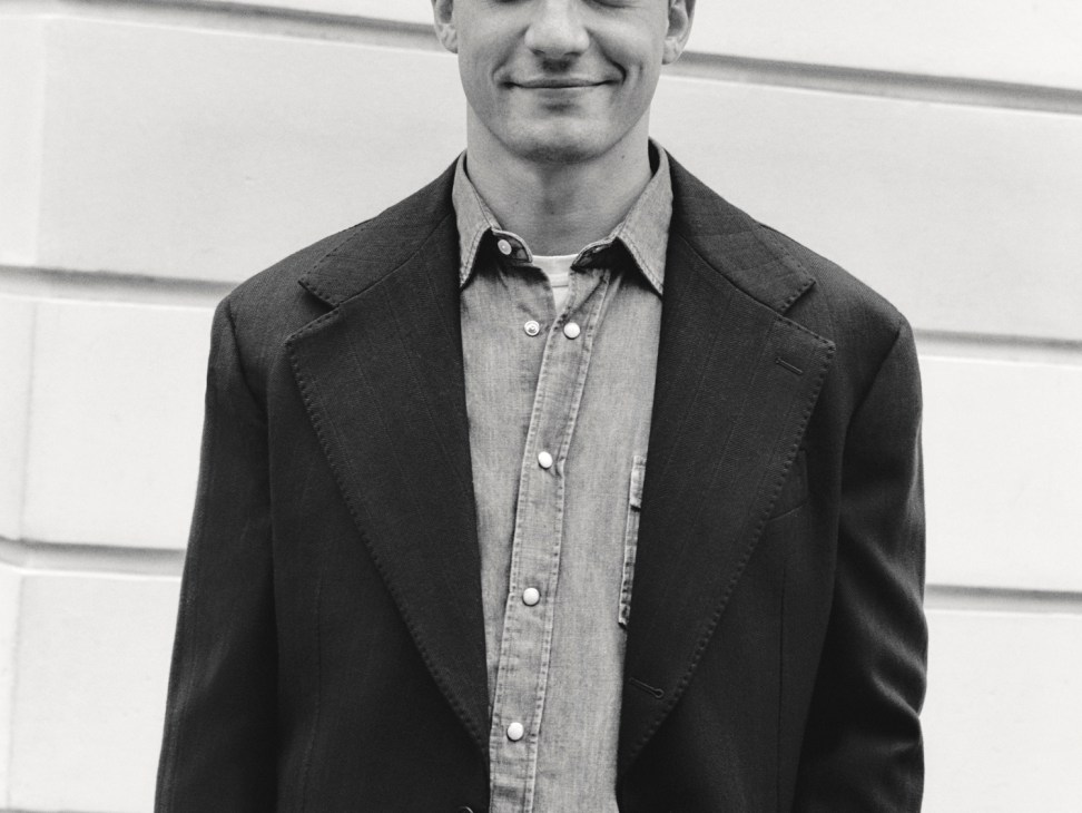 Model wearing dark blazer over light button-up shirt with hands in pockets against white paneled wall