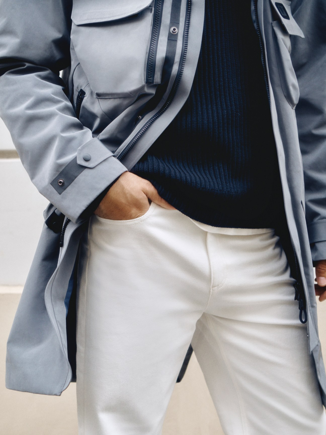 Model in light grey coat over navy ribbed jumper with white trousers, hand in pocket
