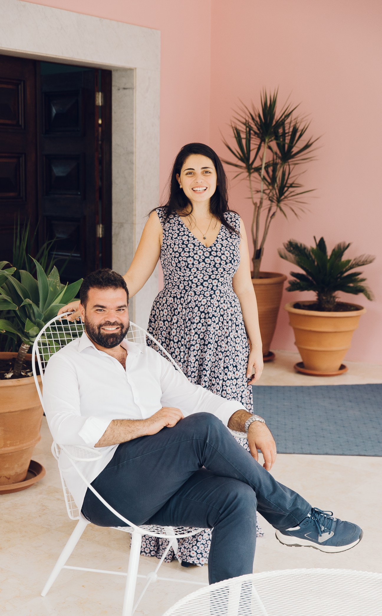 Two people pose together in a minimalist interior with pink walls and potted plants.