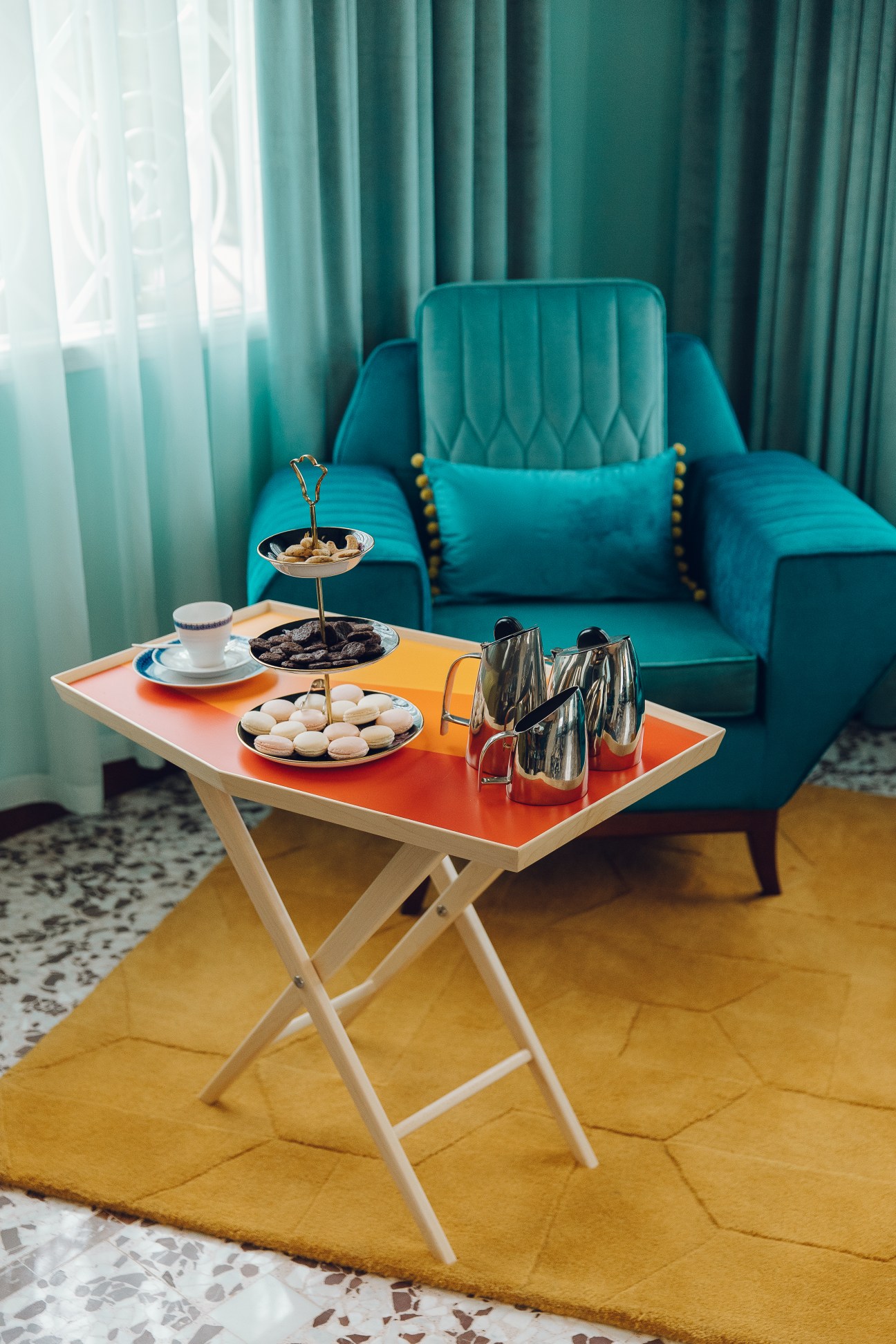 Mid-century modern living room with turquoise velvet chair, orange-topped table with tea service and pastries, teal curtains, and gold accents.