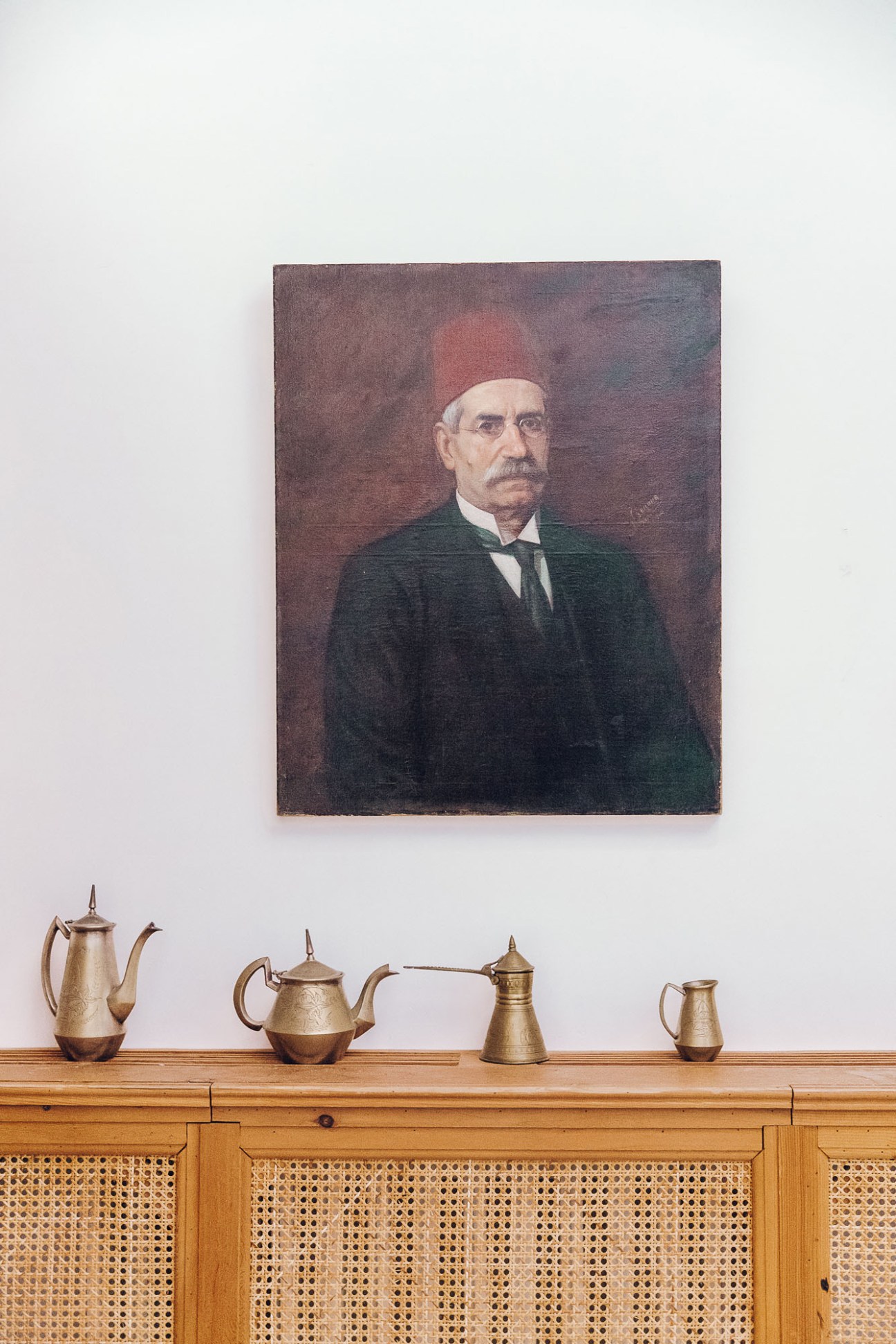 Portrait painting by Habib Srour of a man in a red fez and dark suit hanging above brass vessels on a wooden cabinet.