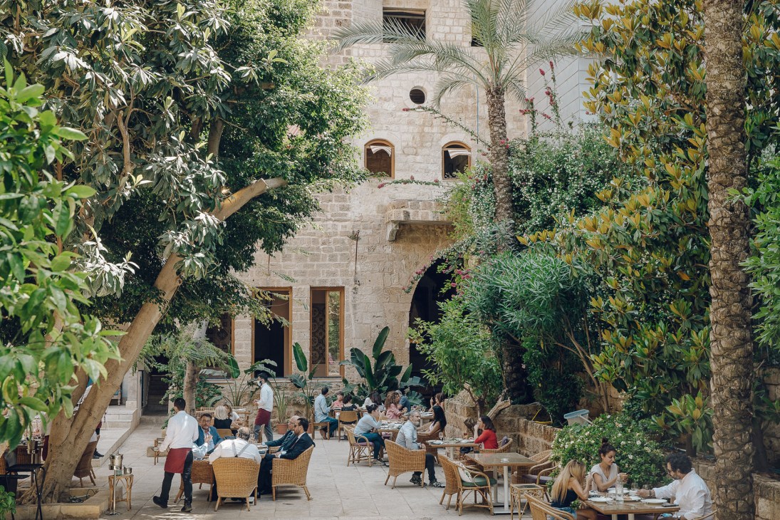 Diners seated at tables in a lush garden courtyard surrounded by greenery with historic stone architecture.
