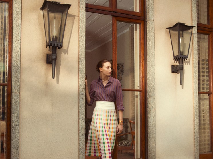Woman in purple striped shirt and colorful checkered skirt standing in doorway of stone building
